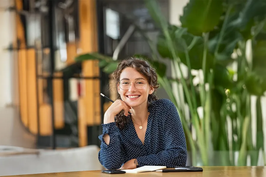Frau mit lockigem Haar und blauem, gepunktetem Oberteil sitzt an einem Holztisch mit Notizbuch, Stift und Smartphone und grünen Pflanzen im Hintergrund