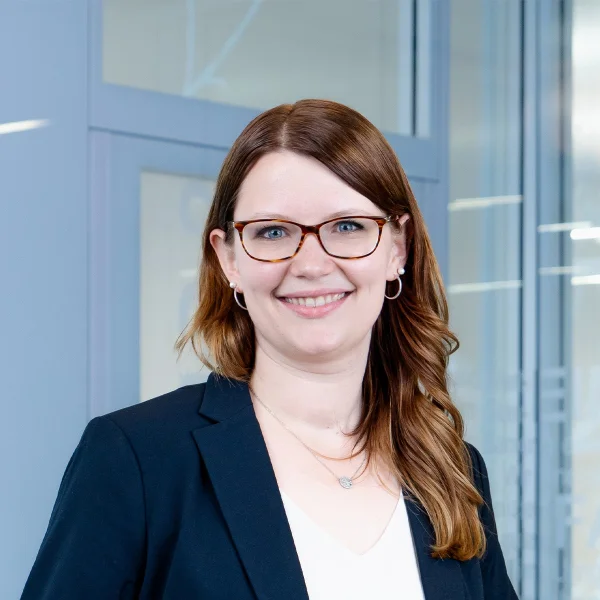 Frau mit langen, gewellten braunen Haaren trägt schwarzen Blazer, weiße Bluse, silberne Ohrringe und zwei Ketten vor Glaswand