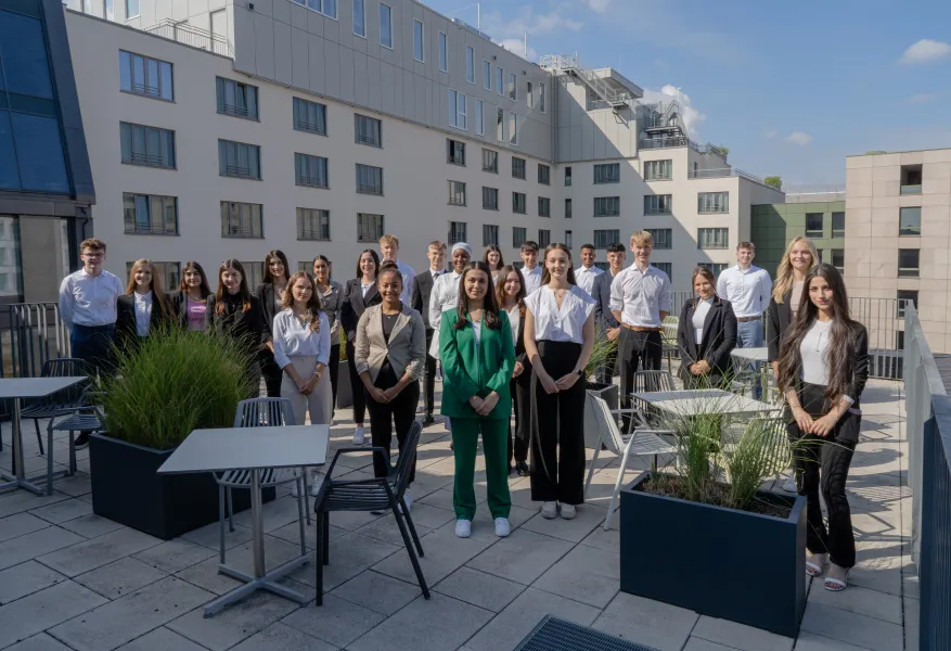 Gruppe von jungen Erwachsenen in Businesskleidung steht auf einer Dachterrasse vor einem modernen Bürogebäude bei sonnigem Wetter