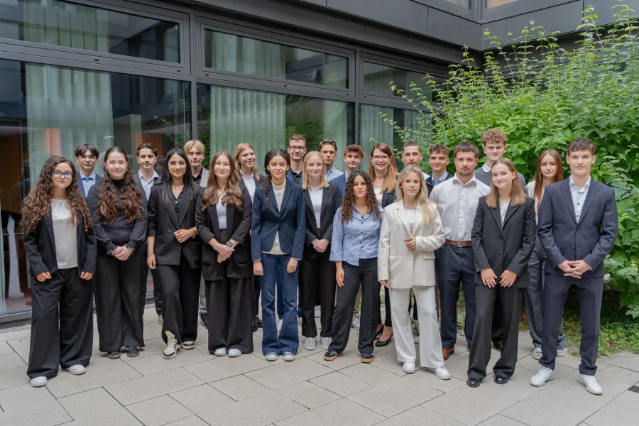 Gruppe junger Erwachsener in formeller Kleidung steht vor einem modernen Gebäude mit großen Fenstern und einem grünen Busch