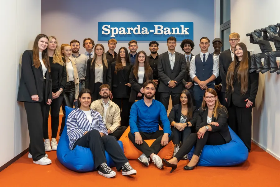 Gruppe junger Erwachsener in Businesskleidung vor Wand mit blauem Schild 'Sparda-Bank', einige sitzen auf bunten Sitzsäcken