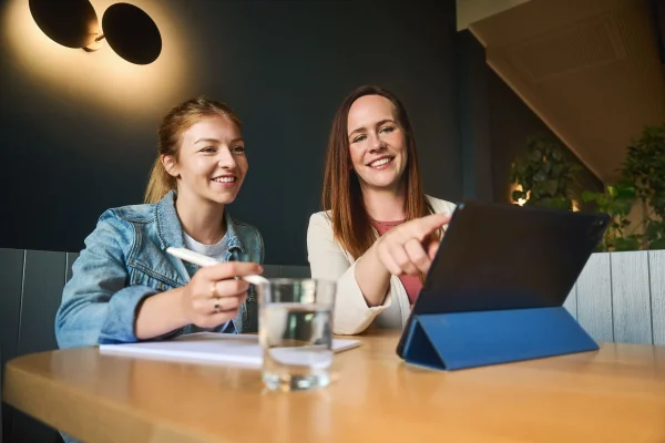Zwei Frauen sitzen an einem Holztisch und schauen auf ein Tablet, eine Frau hält einen Stift, auf dem Tisch steht ein Glas Wasser
