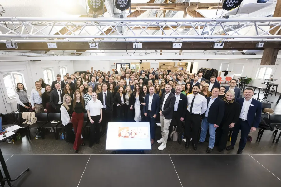 Große Gruppe von Menschen in einem hellen Raum mit Holzbalkendecke, die in Richtung Kamera blickt