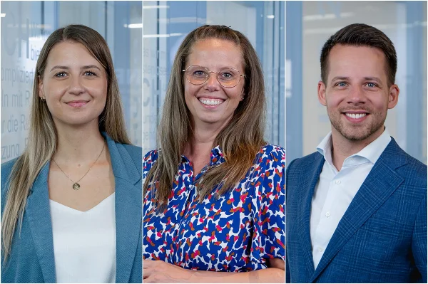 Drei Personen in Businesskleidung vor einer Glaswand mit Text im Hintergrund