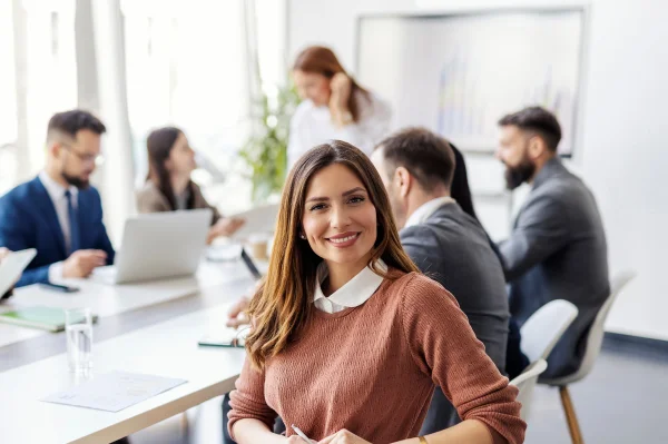 Eine Frau sitzt im Vordergrund. Hinter ihr sind mehrere Menschen an einem Konferenztisch zu sehen.