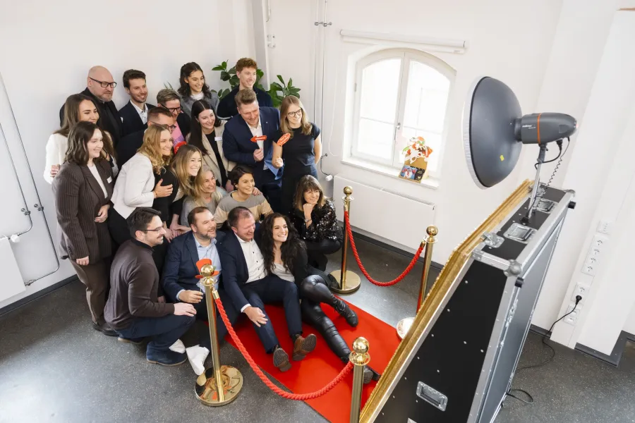 Gruppe von Menschen posiert auf einem roten Teppich vor einer Fotokabine mit Blitzlicht und Absperrung in einem hellen Raum