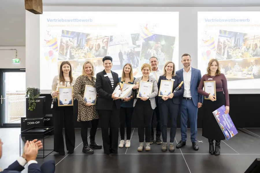 Gruppe von acht Personen steht auf einer Bühne, sieben halten Urkunden, eine hält ein großes Plakat, im Hintergrund zwei Projektionsflächen mit dem Titel 'Vertriebswettbewerb'